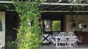 Une terrasse couverte avec un mur en pierre, une table et des chaises blanches, et une plante suspendue.