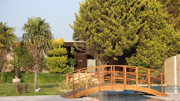 Un pont en bois enjambant une piscine, entouré d’une végétation luxuriante et avec un bâtiment en arrière-plan.