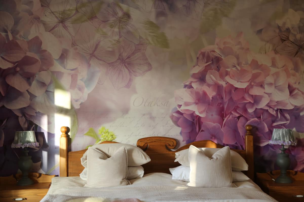 A bedroom with a floral wallpaper, a wooden headboard, two bedside tables with lamps, and a bed with pillows.
