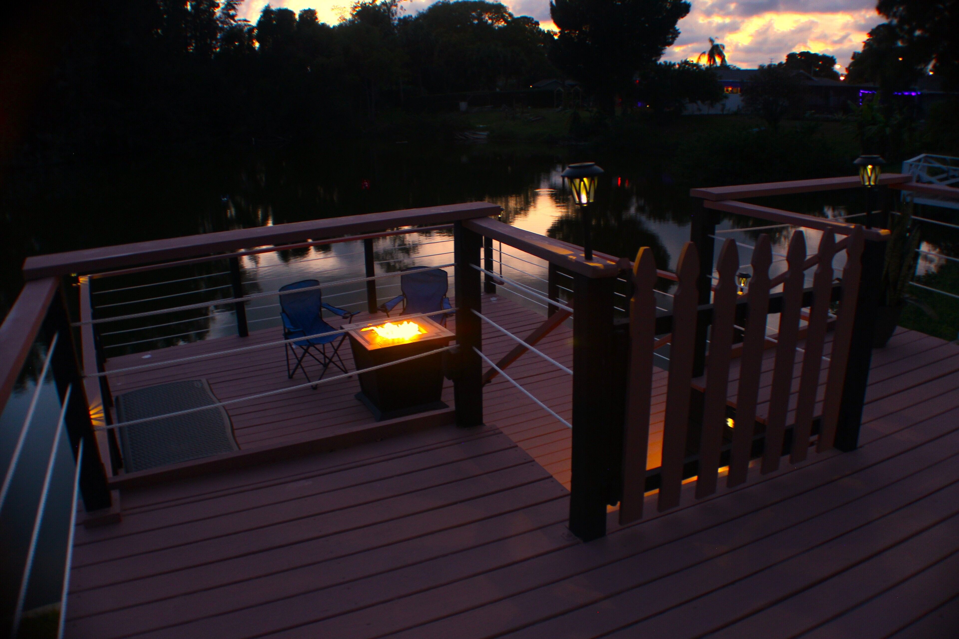 Eine Holzterrasse mit einem Feuerkorb, Stühlen und einem Geländer mit Blick auf ein Gewässer.
