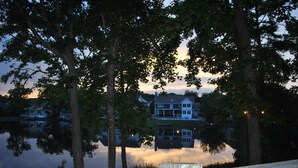 A lakeside view with a wooden deck, trees, and a house reflecting on the water.