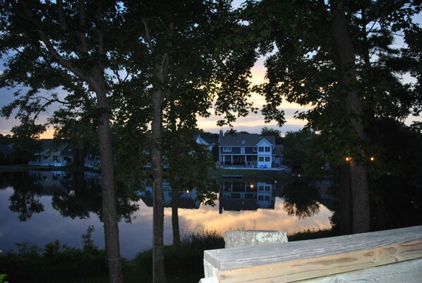 Blick auf einen See mit einer Holzterrasse, Bäumen und einem Haus, das sich im Wasser spiegelt.