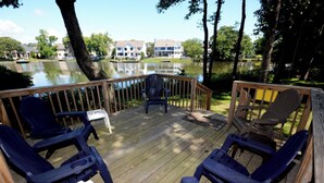 A wooden deck with chairs and a table overlooking a lake, surrounded by trees and houses in the background.