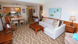 A living room with a sofa, coffee table, and a dining area with chairs and a table. There is a kitchen area with cabinets and a microwave.