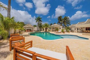 Zona de piscina en un resort con sillones de descanso, palmeras y una cabaña techada de paja.
