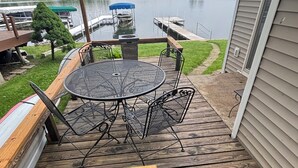 Een houten terras met een ronde metalen tafel en stoelen, uitkijkend op een meer met steigers en boten.