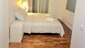 Ein Schlafzimmer mit Bett, Nachttisch, Lampe, einem Fenster mit Vorhängen und Holzboden.