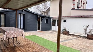 Ein überdachter Patio-Bereich mit Holztisch und -stühlen, einer grauen Garagentür und einem grünen Rasen aus künstlichem Gras.