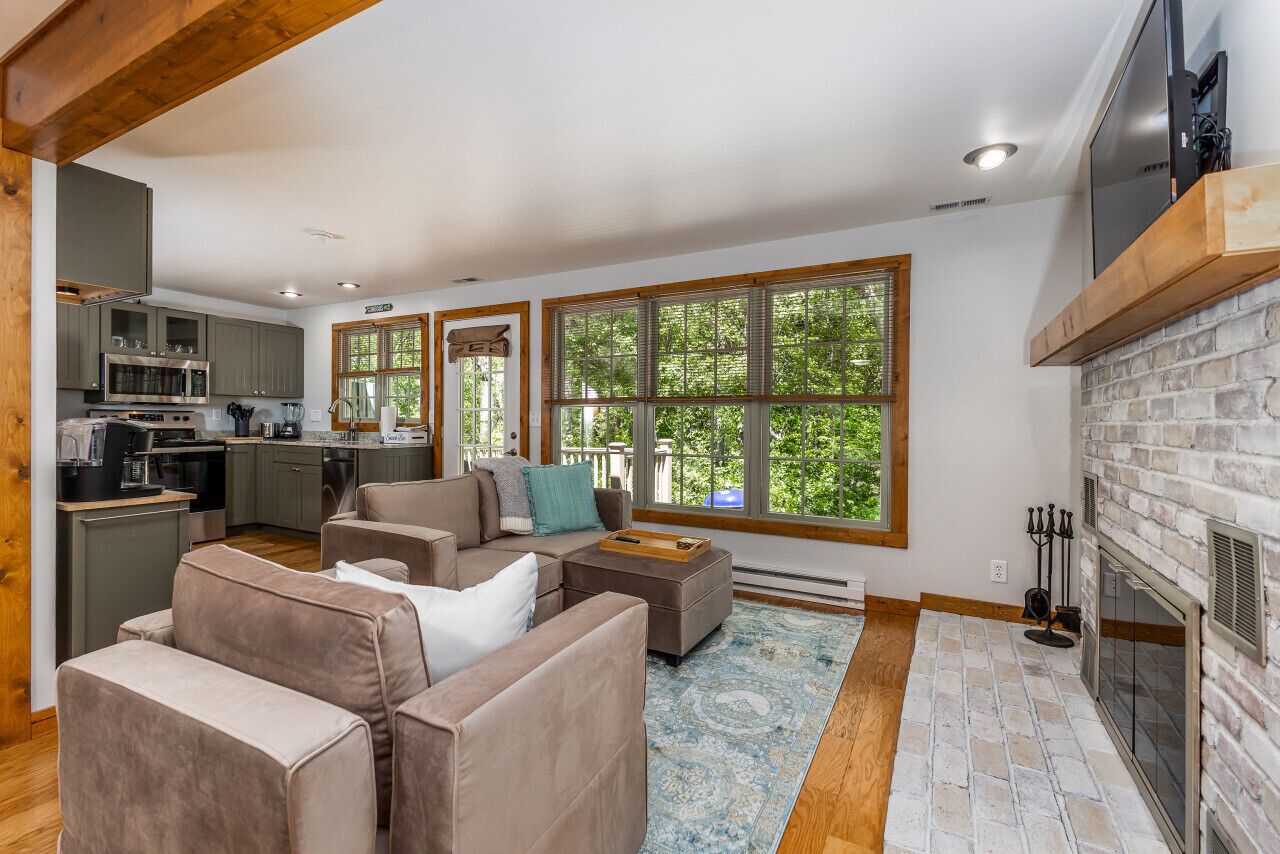 A modern living room with a fireplace, large windows, and a kitchen in the background.