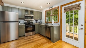 A modern kitchen with stainless steel appliances, wooden flooring, and a view of greenery through a window.