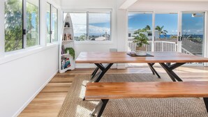 A bright room with a wooden table and benches, large windows, and a view of the ocean. - Coastal Sanctuary with Ocean Views. (Tweed)