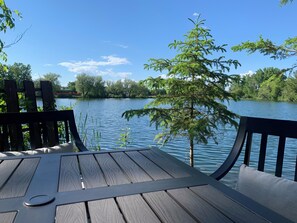 A wooden deck with a view of a calm lake and surrounding trees. - Serene river apartment walking distance to downtown and Heartland Trail! (Park Rapids)