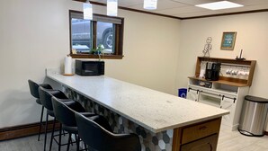 Una cocina con una encimera amplia, taburetes de bar, microondas, cafetera y un cubo de basura.