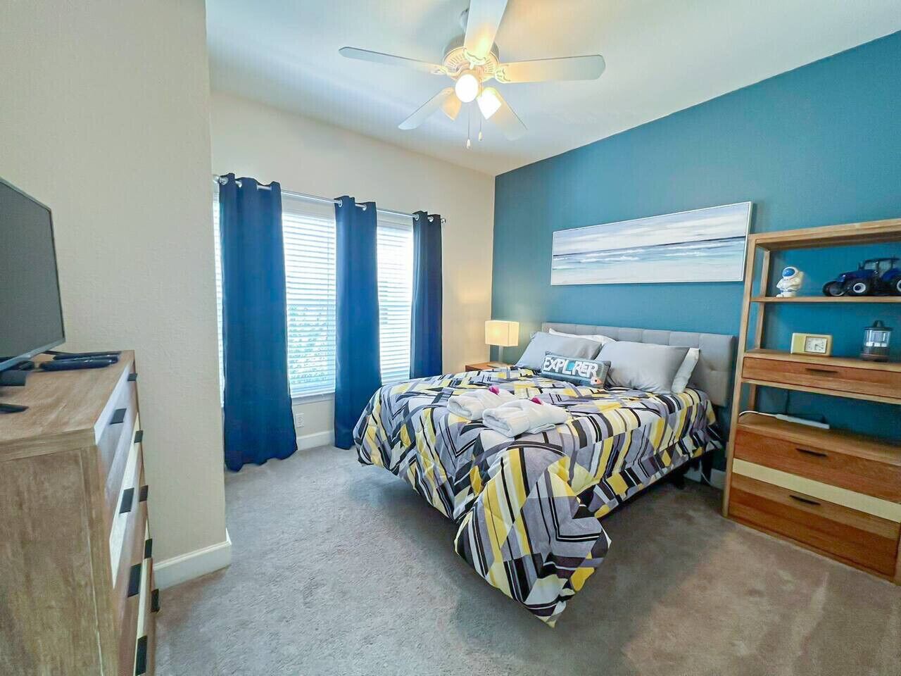 A bedroom with a bed, a ceiling fan, a window with curtains, a dresser, and a shelf with decorative items.