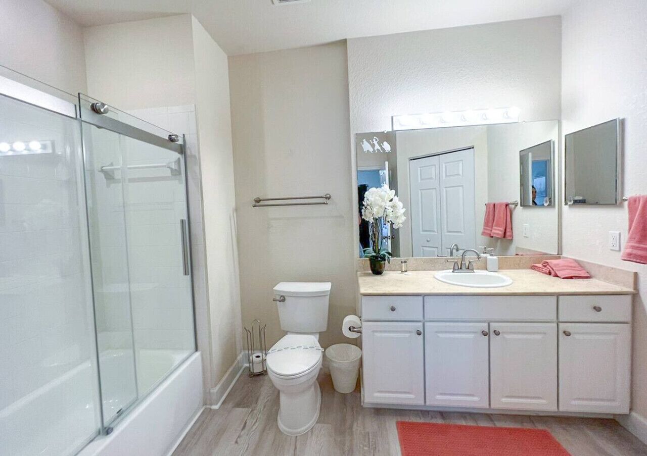 A modern bathroom with a glass shower enclosure, a toilet, a sink with a mirror, and a red bath mat.