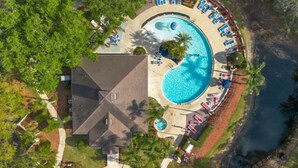 Eine Vogelperspektive zeigt ein Resort mit einem zentralen Schwimmbecken, einem kleineren kreisförmigen Pool und einem Strandbereich mit Liegestühlen.