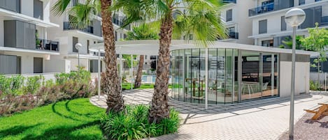 A modern apartment complex with a glass conservatory, palm trees, and a well-maintained garden.