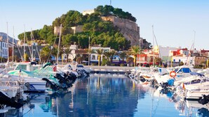 A marina with boats docked in calm waters, a historic castle on a hill in the background, and buildings along the waterfront.