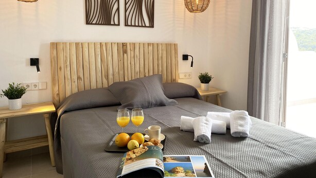 Ein modernes Hotelzimmer mit einem hölzernen Kopfteil, zwei Wandleuchten, einem Nachttisch mit Pflanze und einem großen Bett mit Zeitschrift und Frühstücksartikeln.