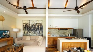 A modern living room with a sofa, coffee table, and artwork. Adjacent is a kitchen with a dining area featuring a fruit bowl and wine glasses. - Villa Chloe by Pertama Stay (Seminyak)