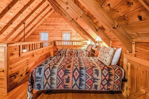 Un dormitorio acogedor en el ático, con techo inclinado de madera, una cama con colcha estampada y dos ventanas con persianas.