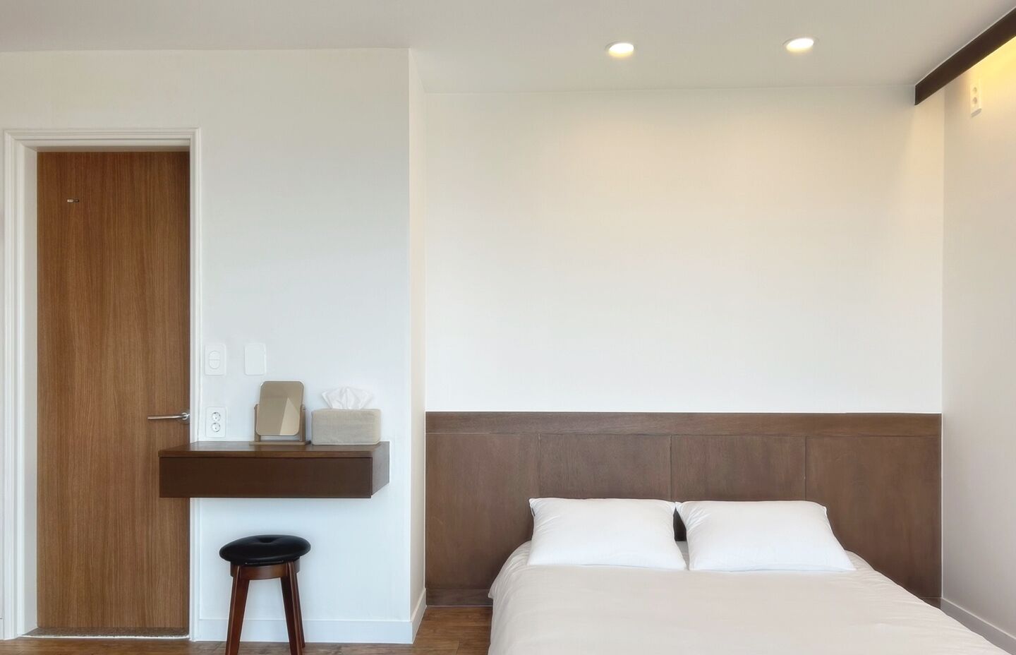 A hotel room with a wooden door, a bedside table with a lamp, a chair, and a bed with white bedding.