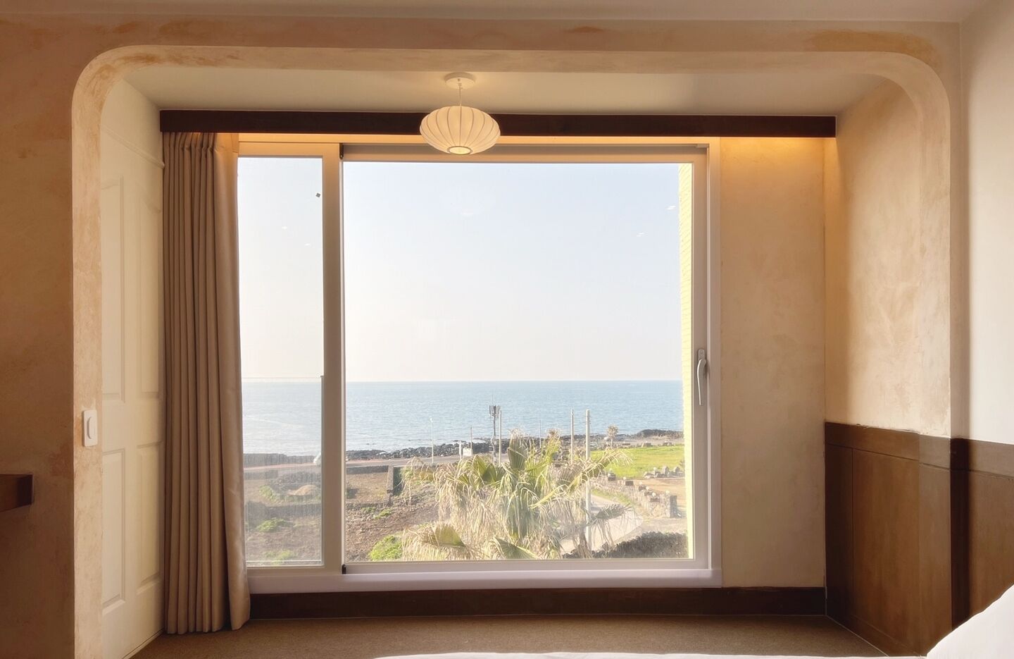 A room with a large window overlooking a coastal landscape, a ceiling light fixture, and a door with a curtain.