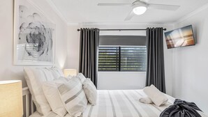 Una habitación de hotel con cama, ventilador de techo, ventana con cortinas, televisor montado en la pared y una obra de arte enmarcada.