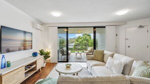 Una sala de estar moderna con un sofá, una mesa de centro y un televisor. Hay una puerta corrediza de vidrio que da a un patio exterior con una mesa y sillas.