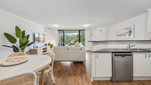 Una cocina moderna con armarios blancos, lavavajillas y un comedor con mesa redonda y sillas de mimbre.
