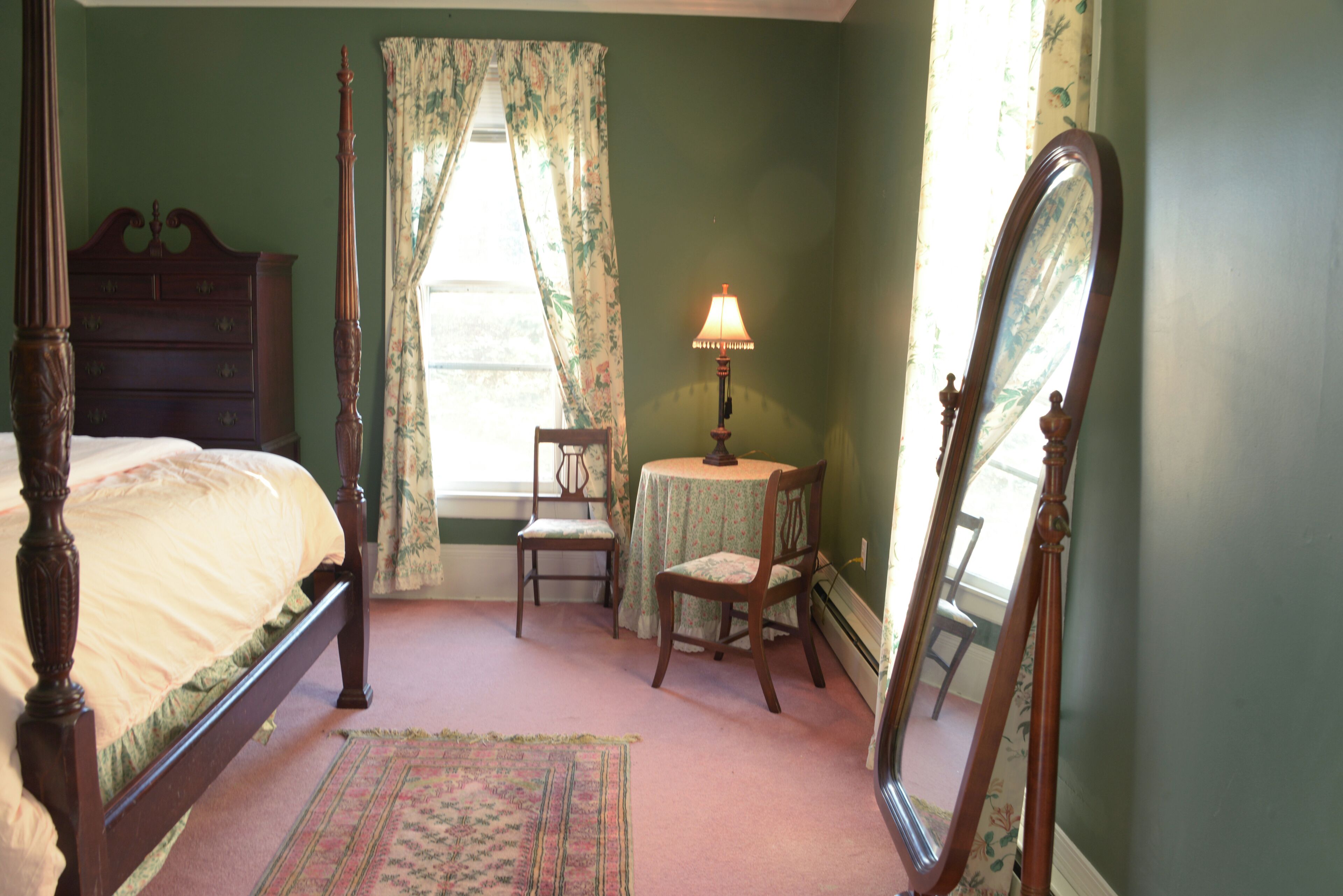 A bedroom with a four-poster bed, a round table with a lamp, a chair, and a full-length mirror.