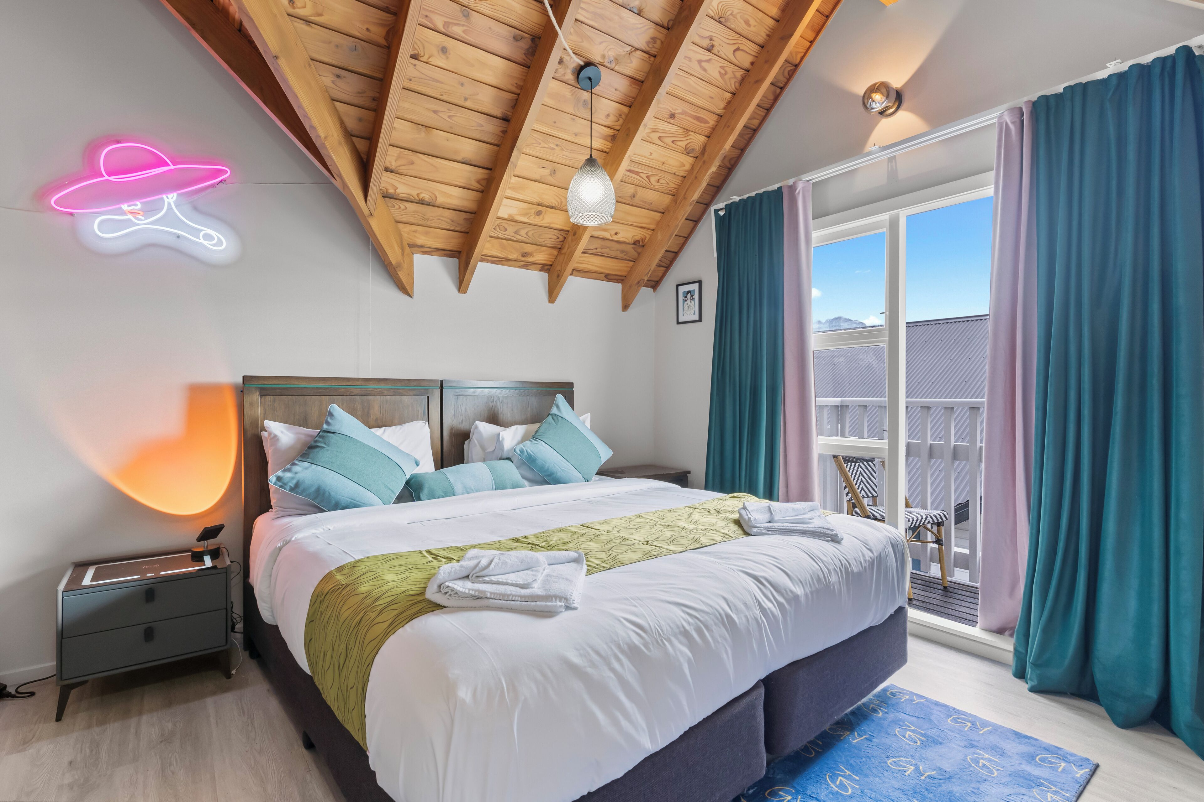 Un dormitorio con techo de madera, cama con ropa de cama blanca y azul, un letrero luminoso con forma de sombrero y un balcón con cortinas.
