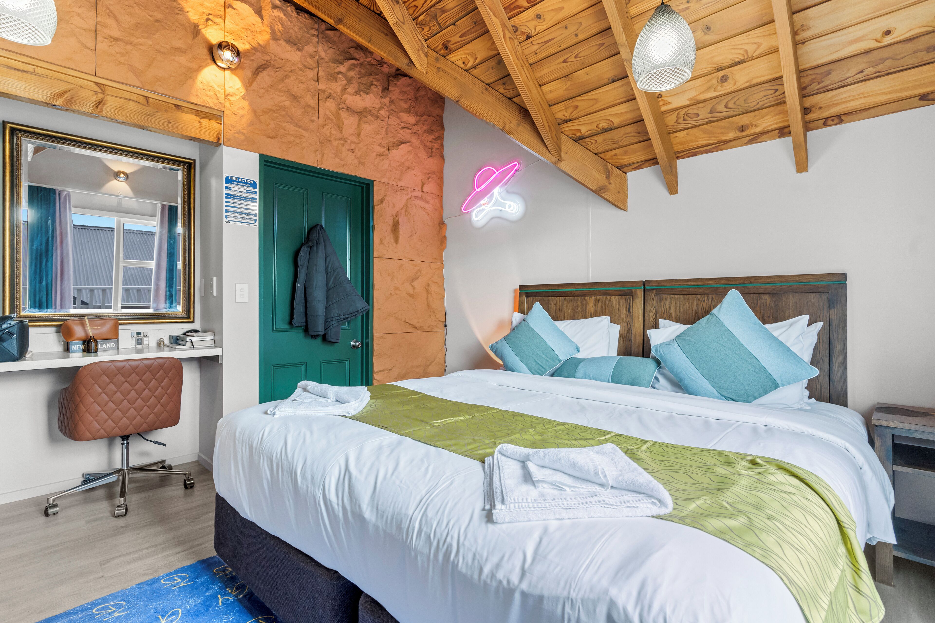 A bedroom with a wooden ceiling, a bed with white and green bedding, a desk with a chair, a neon sign, and a green door.