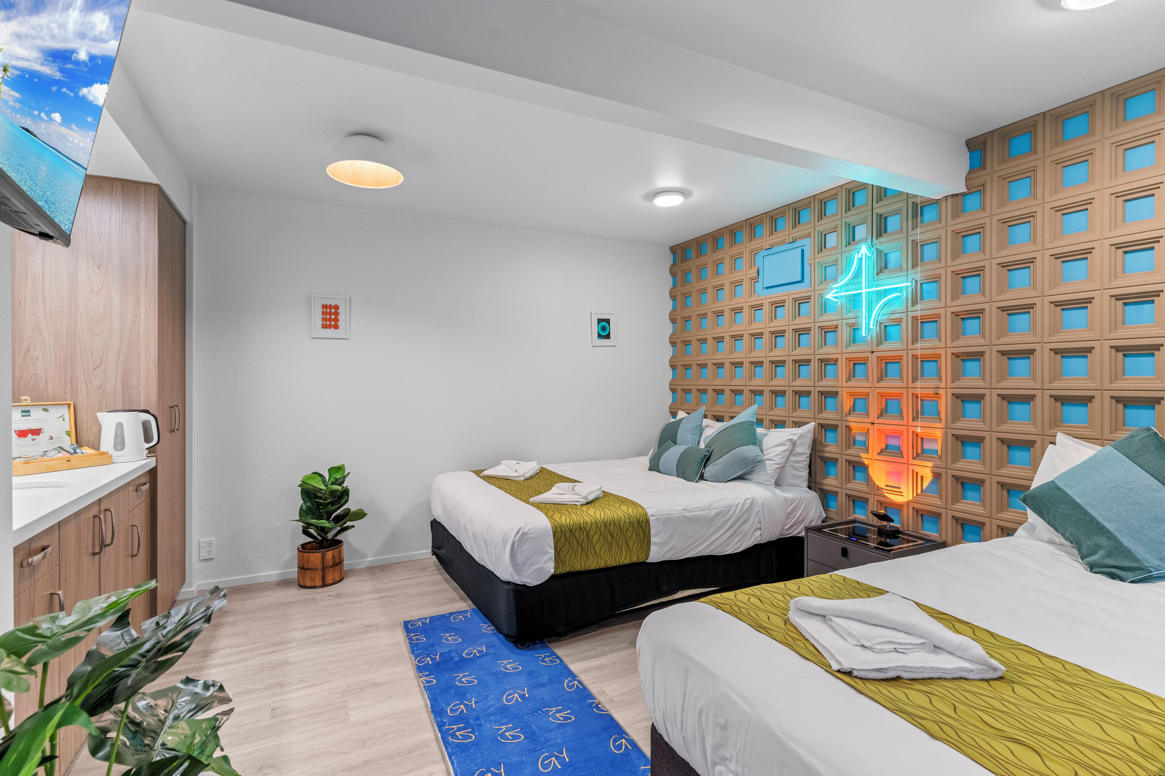 Ein modernes Hotelzimmer mit zwei Betten, einem Holz-Kopfteil, einem Neonlichtschild und einer kleinen Pflanze.