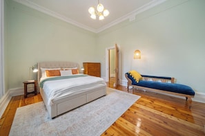 Quarto com cama, mesa de cabeceira, abajur, banco e porta para outro cômodo.