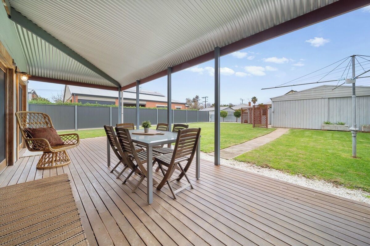 A covered outdoor patio with a table and chairs, a wicker chair, and a view of a backyard with a fence and a building.