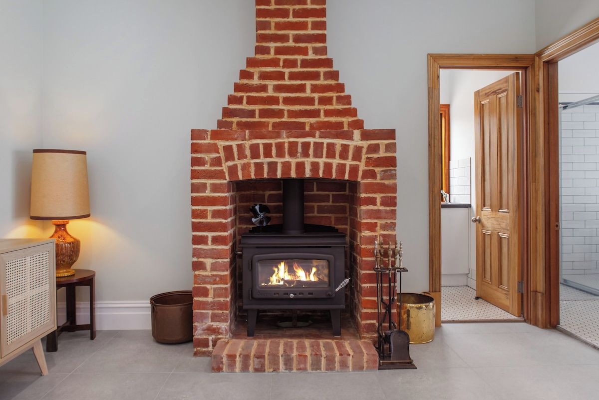 Een metselwerk open haard met een moderne houtkachel, een lamp op een bijzettafeltje en een houten deur op de achtergrond.