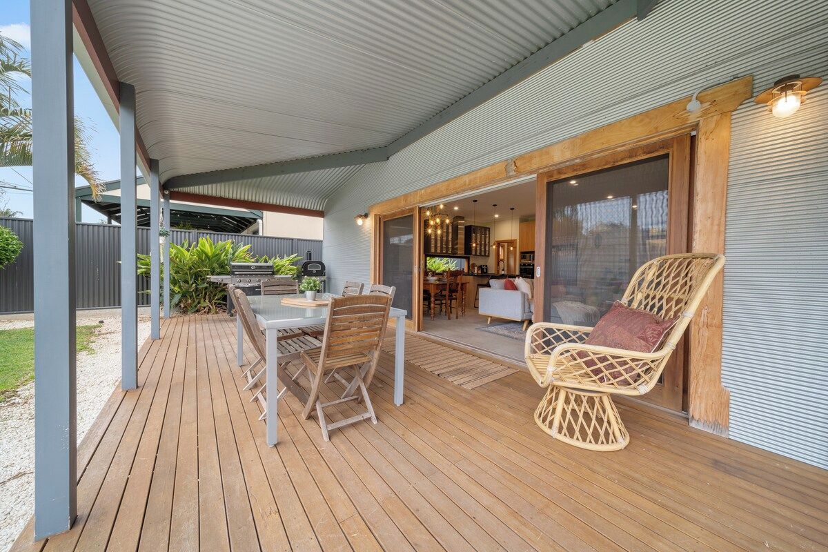 A covered outdoor patio with a wooden deck, a dining table with chairs, and a wicker chair.