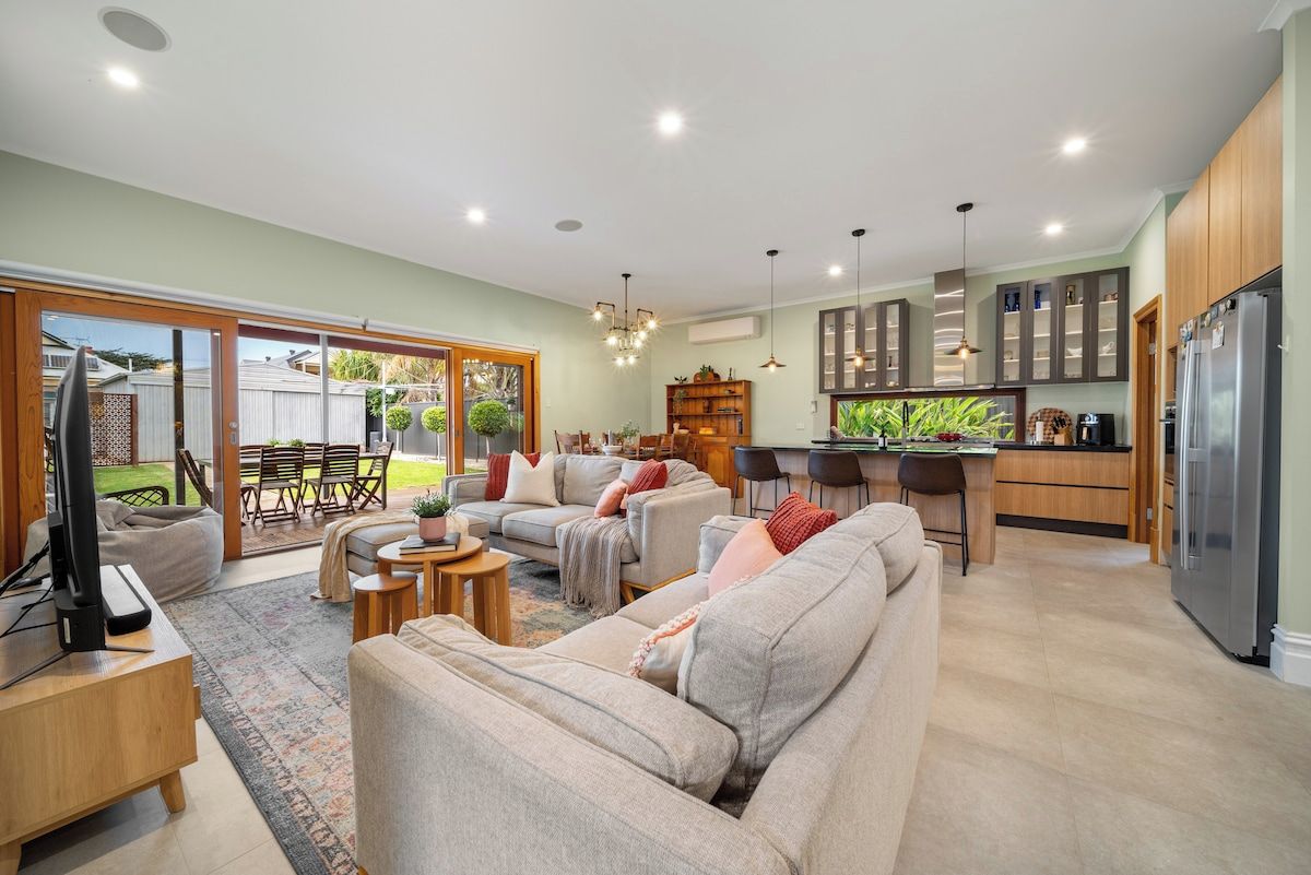 Spacious open-plan living with plush sofas, a modern kitchen, and seamless indoor-outdoor flow to a lush dining area. Perfect for relaxing and entertaining.