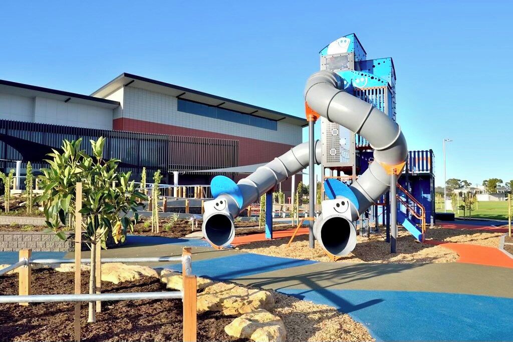 A playground with slides and climbing structures, a building with a red and white facade, and a clear sky.