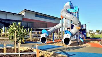 A playground with slides and climbing structures, a building with a red and white facade, and a clear sky.
