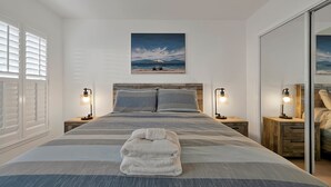 Un lit bien fait, recouvert d’une couvre-lit à rayures, avec deux lampes de chevet, une serviette pliée et un tableau représentant une scène de plage au-dessus du lit.