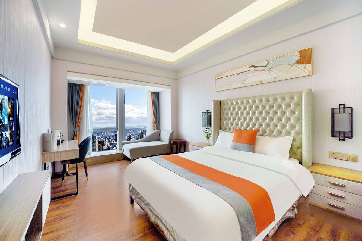 A hotel room with a large bed, a desk, a chair, and a view of the city through the window.