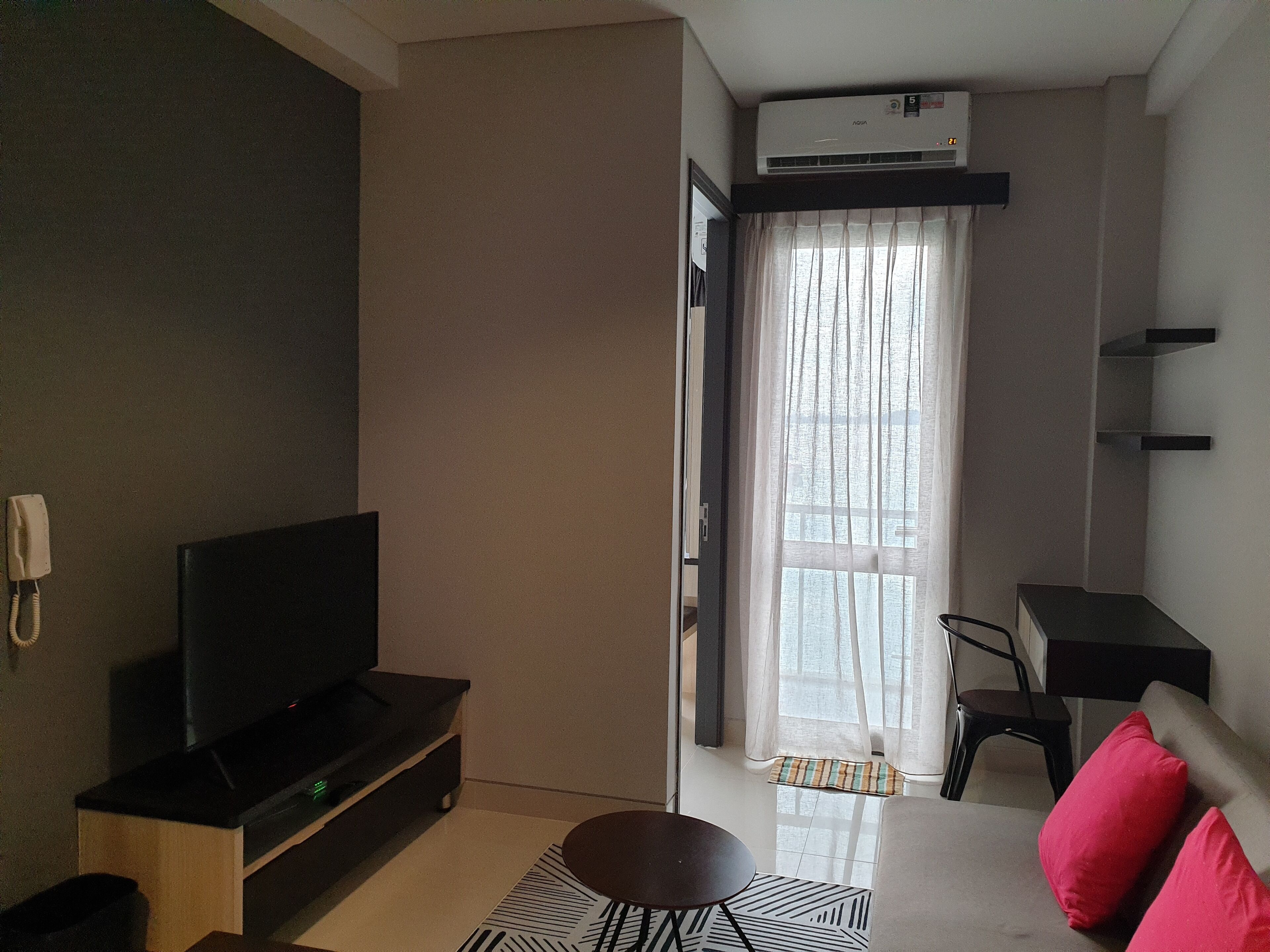 A modern living room with a flat-screen TV on a wooden stand, a small round table, and a sofa with red cushions. There is a window with sheer curtains and an air conditioning unit mounted above it.