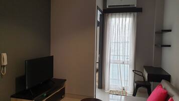 A modern living room with a flat-screen TV on a wooden stand, a small round table, and a sofa with red cushions. There is a window with sheer curtains and an air conditioning unit mounted above it.