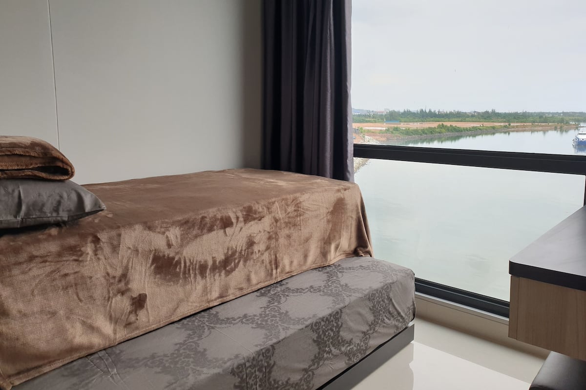 Ein Schlafzimmer mit einem Bett, einem Fenster mit Blick auf ein Gewässer und die Ferne sowie einem kleinen Tisch neben dem Bett.