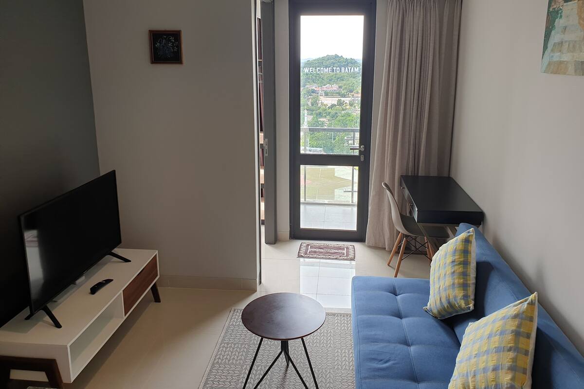 Ein modernes Wohnzimmer mit Fernseher, einem blauen Sofa mit gemusterten Kissen, einem kleinen runden Tisch und Blick durch eine Glastür auf einen Balkon.