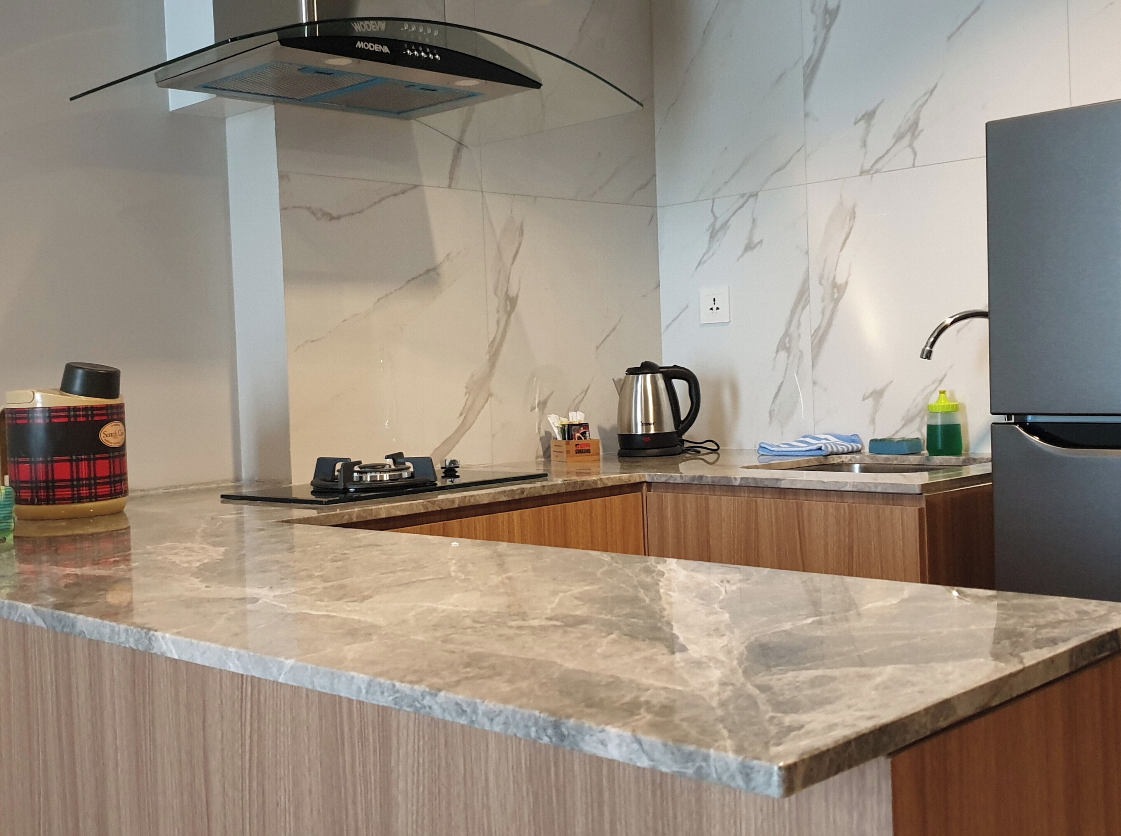 A modern kitchen with a marble countertop, a gas stove, a kettle, and a refrigerator.