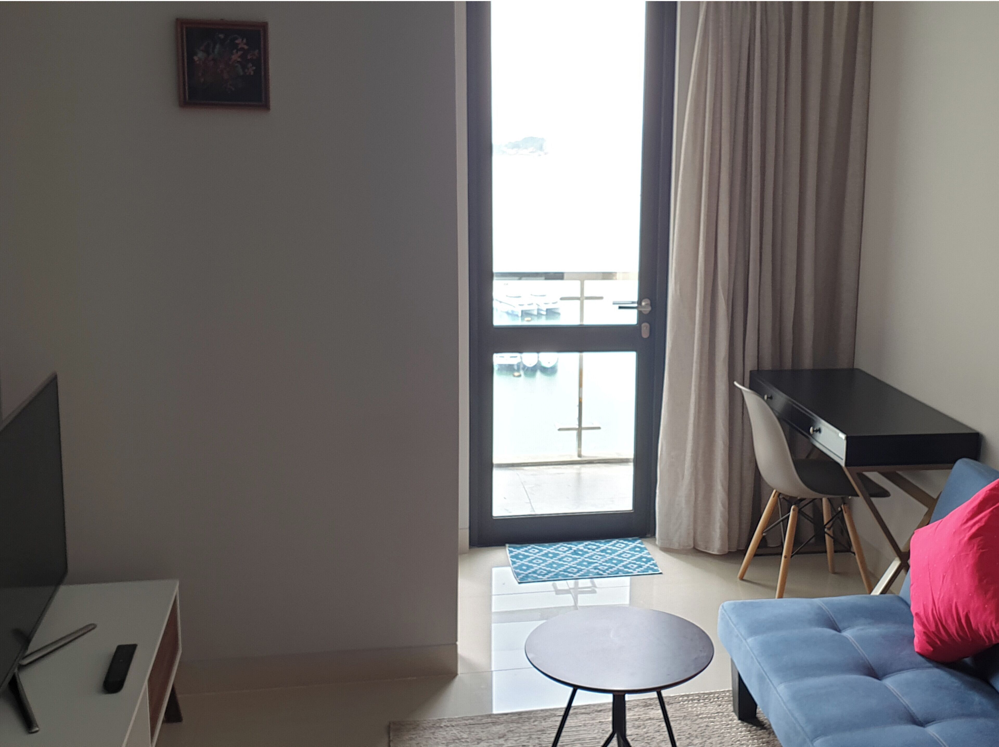 A modern living room with a sofa, a small round table, a desk, and a view of a balcony with a railing.