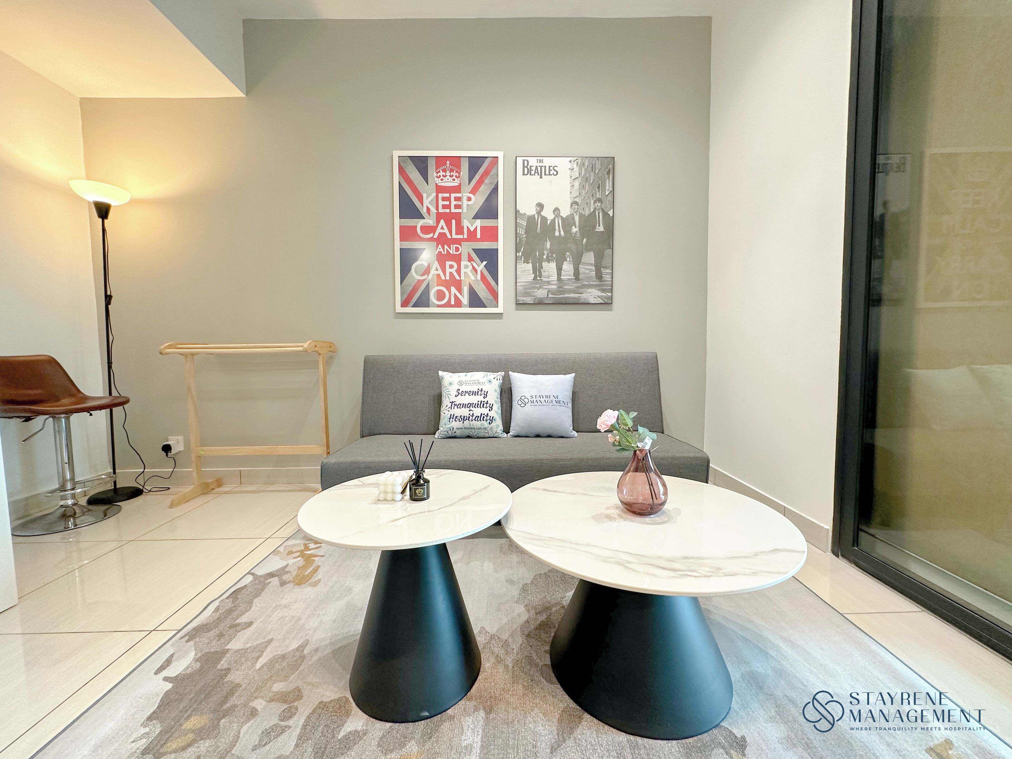 A modern living area with a grey sofa, two round tables, and a lamp. There are posters on the wall, one of which says "KEEP CALM AND CARRY ON".